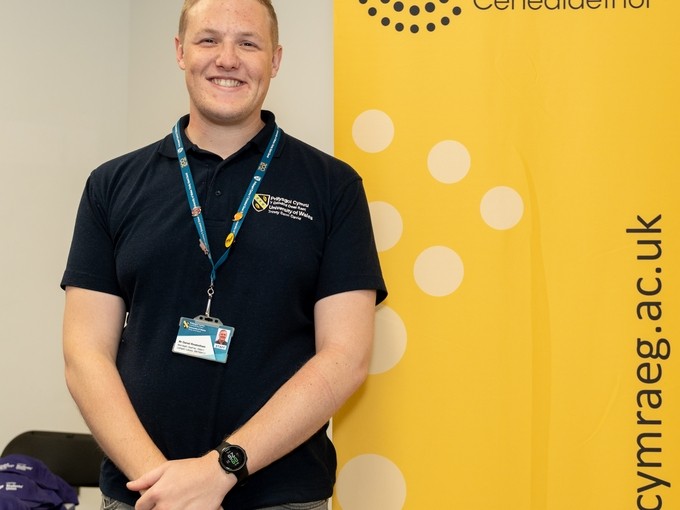 Aelod o staff yn sefyll o flaen baner felen y Coleg Cymraeg