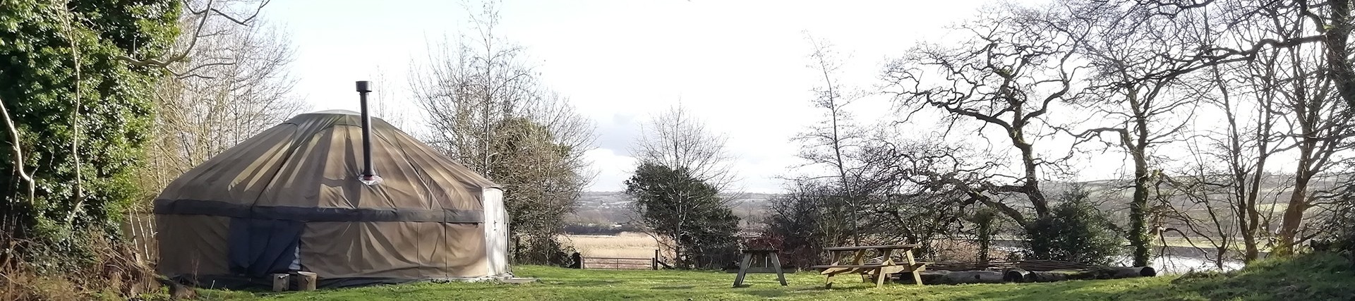 A large canvas yurt in a small field; the yurt has a metal chimney; there is a seating area outside with logs and picnic benches; river meadows and the Towy itself are visible in the background.