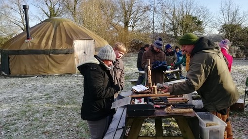 People in a wintry field inspect wood carving equipment set out on a picnic table; a yurt is visible in the background.