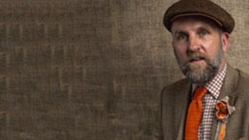 Marc Rees wears an orange tie and orange-patterned cloth flower over his breast pocket; the light-brown shade of his wool blazer matches the wall behind him; he wears a traditional brown flat cap.
