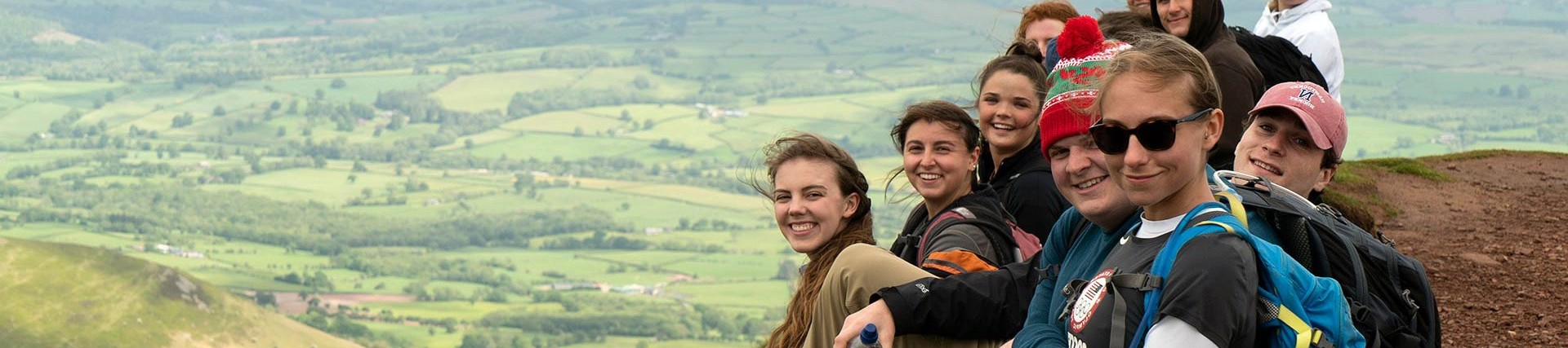 A small group of smiling young people sit on the flank of Pen Y Fan, the green fields of Powys far below them.