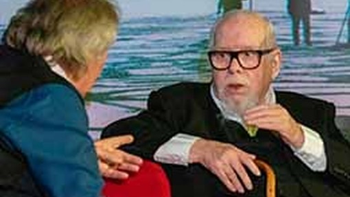 Sir Peter Blake sits in a chair during an interview and raises a hand in emphasis.