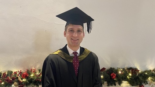 Brandon Gorman yn gwenu wrth wisgo het a gŵn academaidd sydd â chwfl aur.