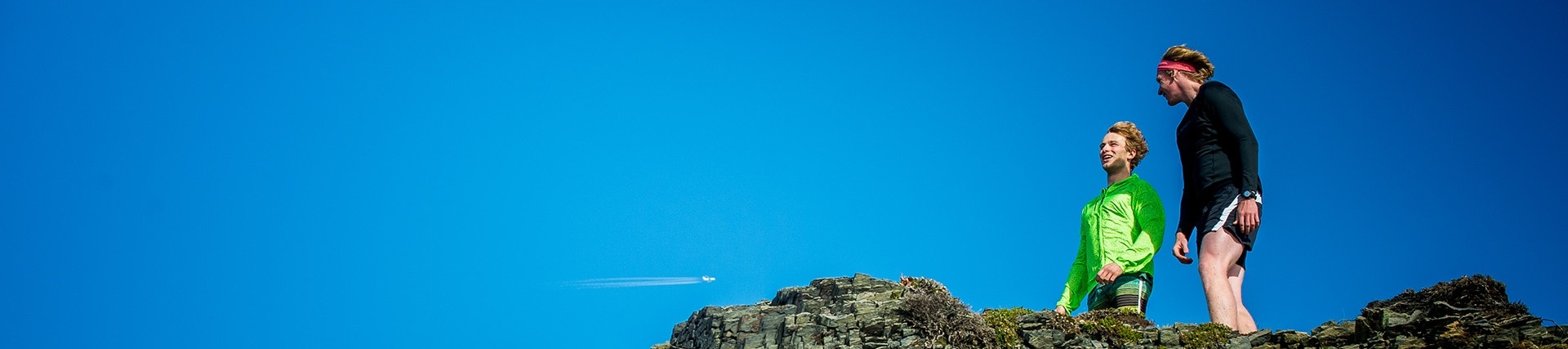 Two young men in running gear stand on an outcrop of rock below a bright blue sky.