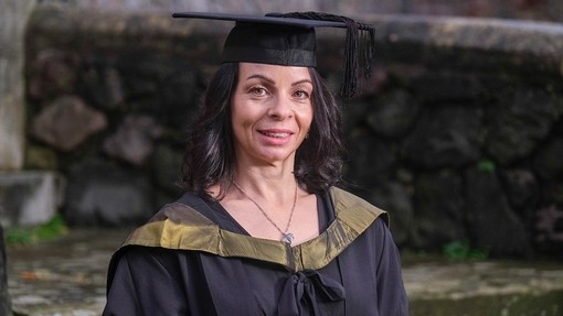 Mae Emma Williams yn gwenu fymryn wrth wisgo het a gŵn academaidd sydd â chwfl aur.