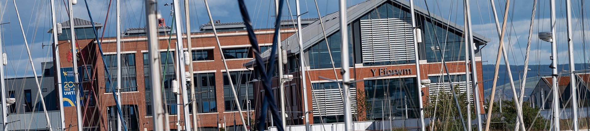 Y Fforwm Library, a large building with pillars of red brick and much glass. The masts of yachts are visible in the foreground.